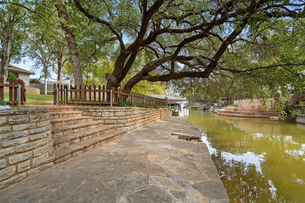 Waterfront hardscape featuring natural stone paving, terraced stone retaining walls, integrated stone stairs, and wood-slat fencing beneath mature canopy trees