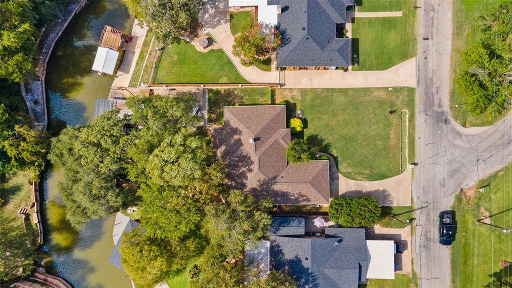Aerial perspective featuring a waterfront property with a concrete driveway, multi-gabled shingle roof, mature trees, and boat dock