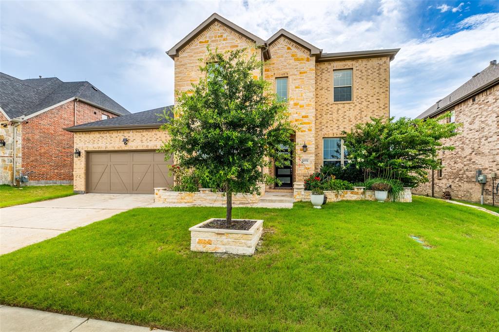 Two-story residence featuring a multi-toned brick facade, gabled roofline, integrated two-car garage with wood-finish doors, and stone-accented landscaping planters