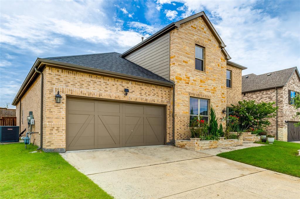 Two-story residence featuring a tan masonry facade, decorative siding accents, a double-car garage with cross-buck detailing, and a stone planter bed