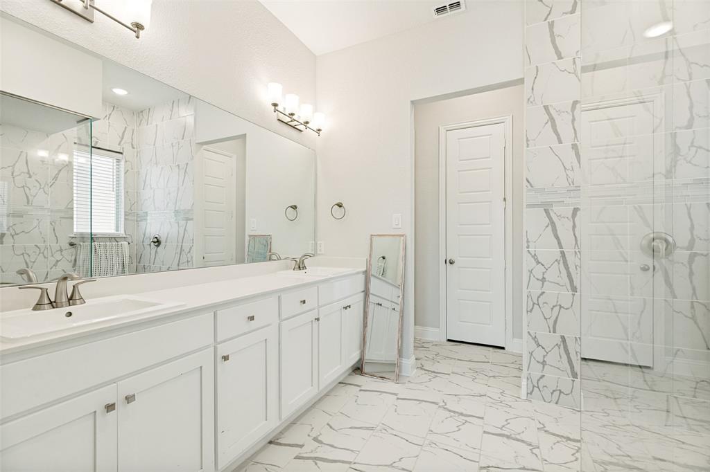 Bathroom featuring a dual-sink vanity with white cabinetry, full-width wall mirror, large-format veined tile flooring, and a walk-in shower with floor-to-ceiling tile surround