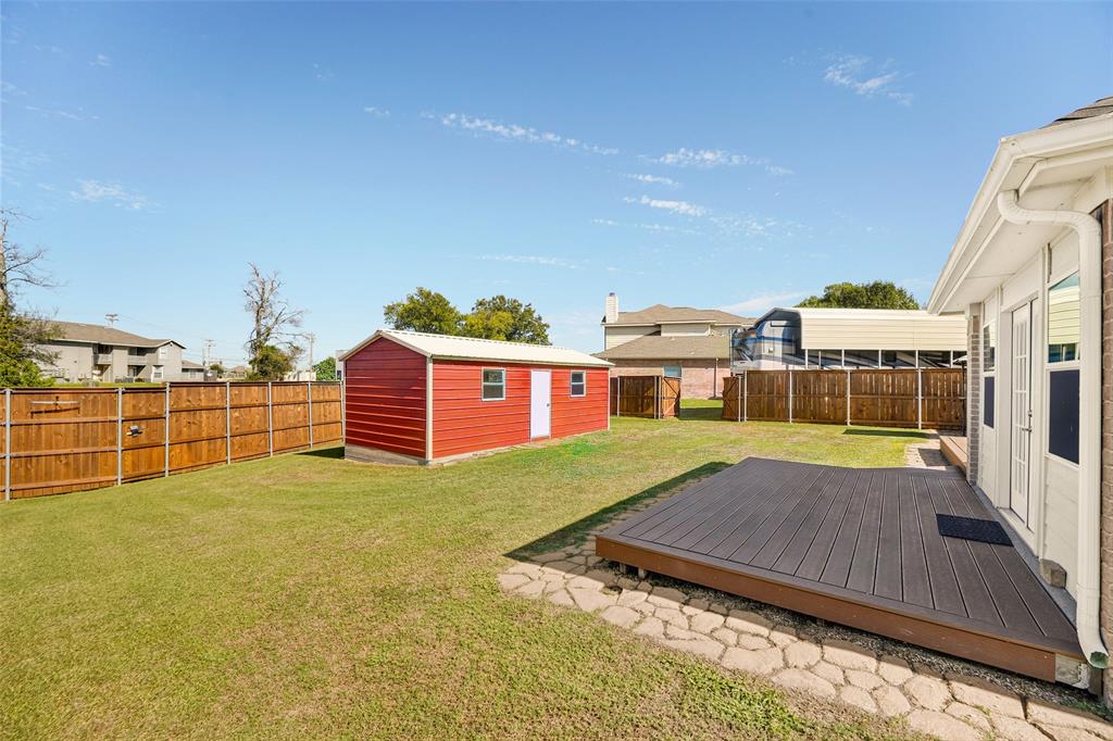 Rear exterior featuring a TREX deck, stone-style paver border, perimeter wood fencing, and a detached metal workshop.  In the background you can see part of the carport and one set of double gates.