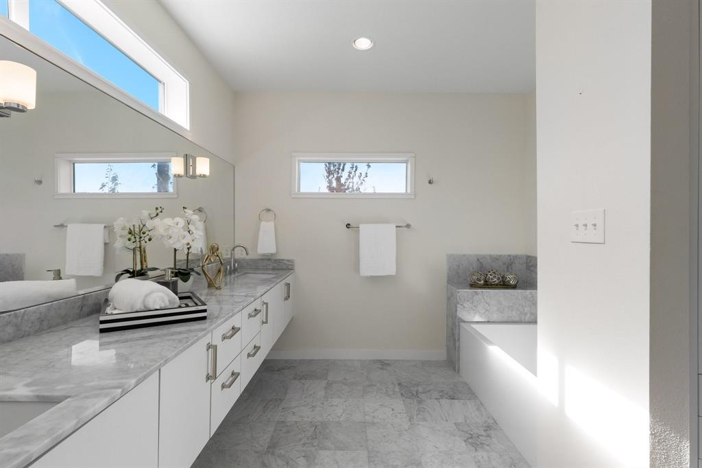 Floating vanity with stone countertops and dual undermount sinks, stone-tiled flooring, stone-surround soaking tub, and clerestory windows