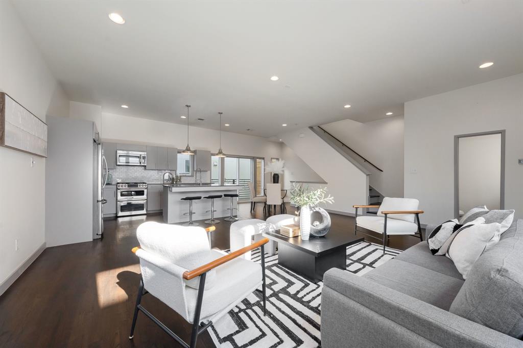 Open-concept living space featuring wood-finish flooring, recessed lighting, stainless steel appliances, a kitchen island with stone countertops, and a staircase with a dark metal railing
