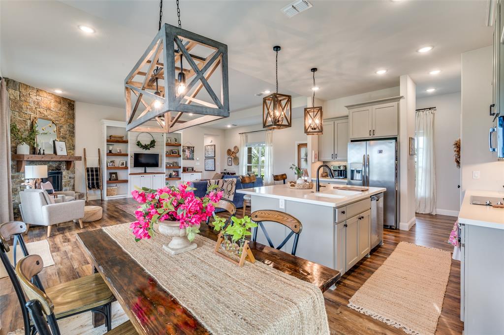 Open-concept area featuring stone-clad fireplace, wood-finish flooring, central island with dual-basin sink, built-in cabinetry, and geometric lantern-style pendant lighting