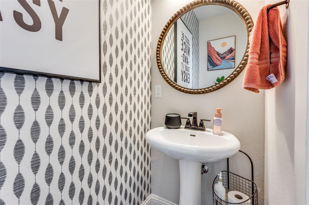 Pedestal sink with a dark metal widespread faucet, patterned wallpaper, and a circular beaded-rim mirror