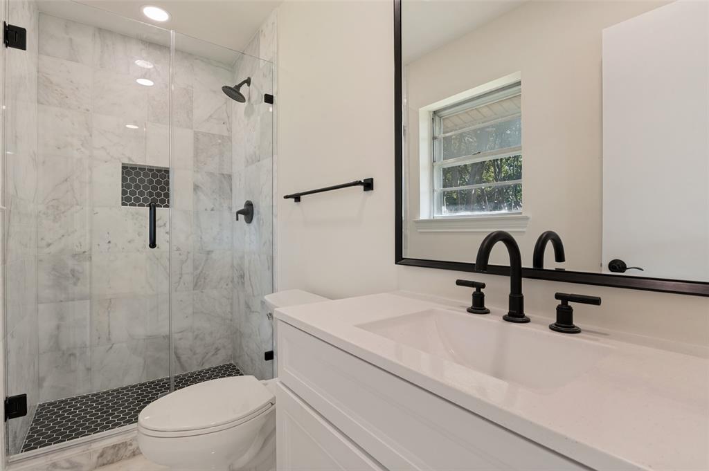 Walk-in shower with stone-tile surround, glass enclosure, and black matte fixtures, paired with a white vanity featuring a quartz countertop and black-framed mirror