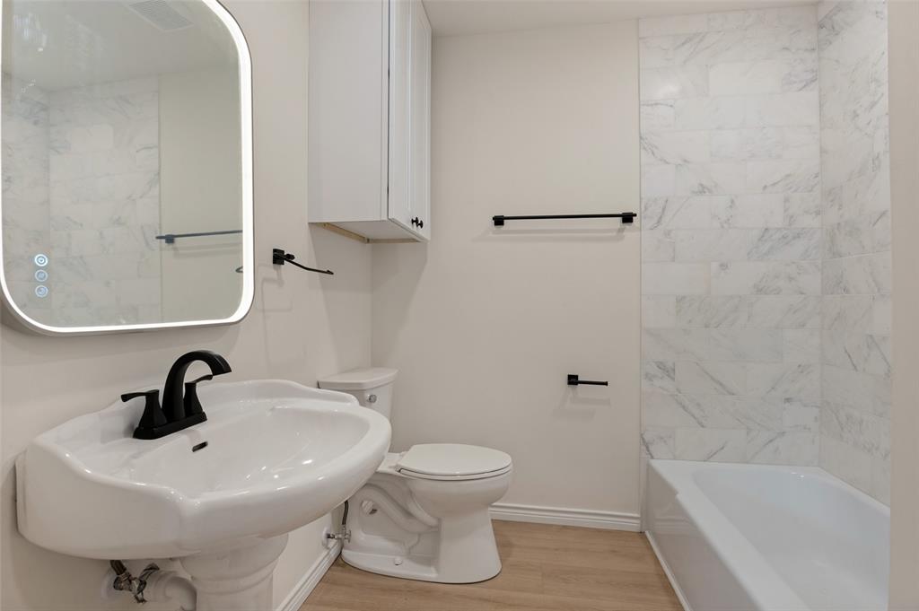 Bathroom featuring a pedestal sink with matte black faucet, wall-mounted backlit mirror, built-in cabinetry, and a tub with stone-finish tile surround