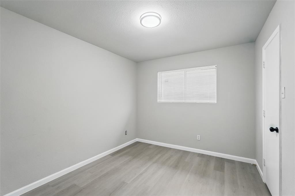 Neutral interior space featuring wood-finish flooring, light gray walls, a flush-mount ceiling light, a horizontal window with blinds, and a paneled interior door with matte black hardware