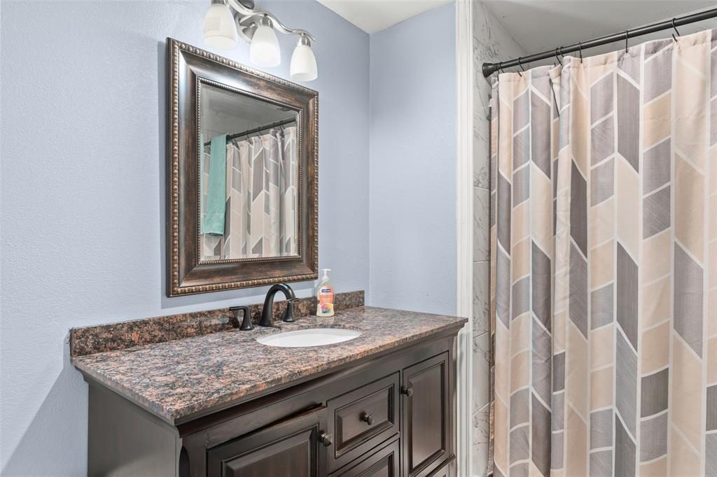 Dark wood vanity featuring a stone countertop, an integrated oval sink, a wall-mounted framed mirror, a three-light fixture, and matte black hardware