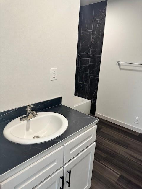 Bathroom featuring white cabinetry with matte black hardware, dark stone-finish countertop with an undermount sink, wood-finish flooring, and a tub enclosure accented with floor-to-ceiling dark stone-finish wall tile