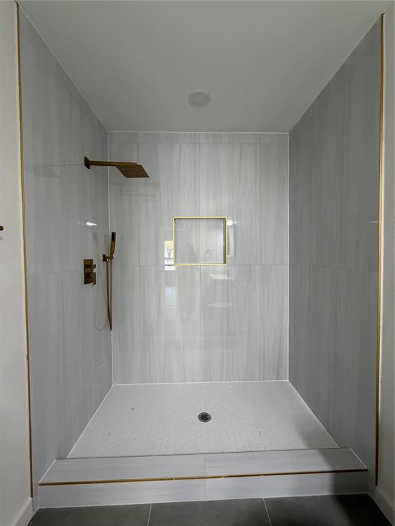 Walk-in shower featuring floor-to-ceiling neutral tile, rainfall showerhead with secondary handheld sprayer, gold-toned hardware, and a built-in tiled wall niche