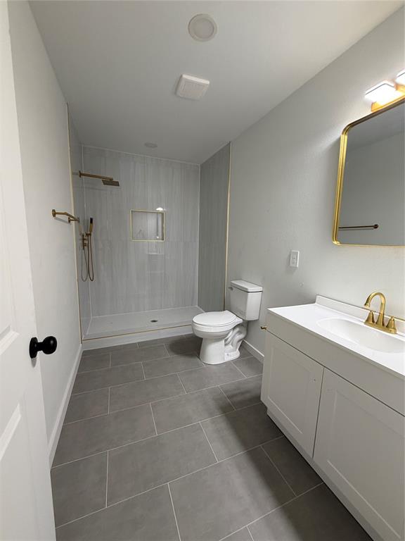 Bathroom featuring a walk-in shower with vertical tile surround and built-in recessed niche, white vanity cabinet with a integrated sink, gold-tone plumbing fixtures, and large-format neutral floor tile