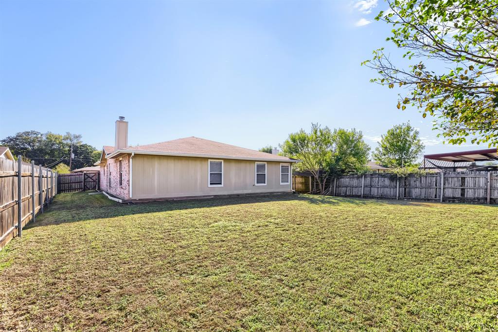 Expansive backyard featuring a grass lawn, full-perimeter wood fencing, a masonry chimney, and a metal-roofed carport structure