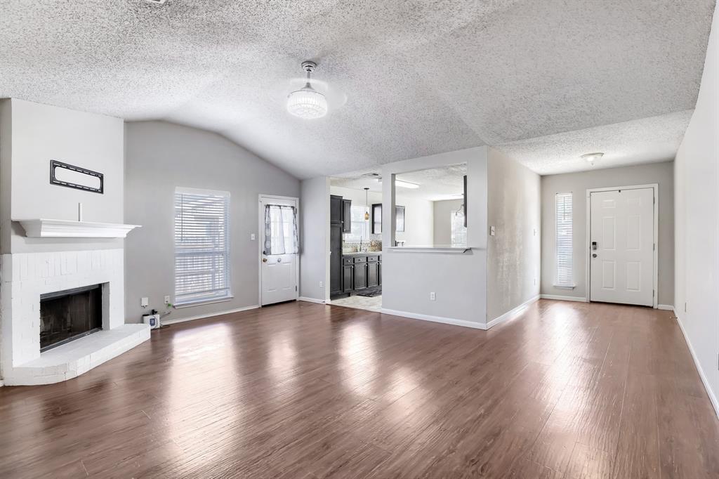 Vaulted ceiling featuring a fireplace with a white brick surround and mantel, wood-finish flooring, and natural light provided by multiple windows and glass-paneled exterior doors