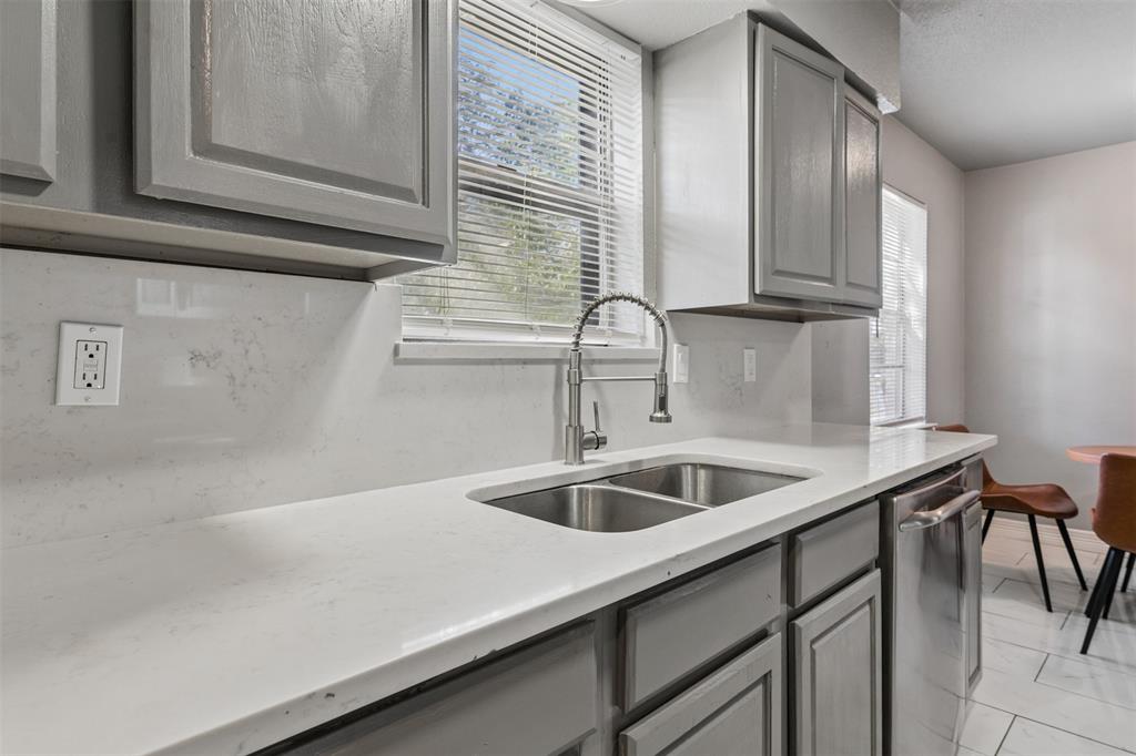 Kitchen featuring grey shaker-style cabinetry, white countertops, a full-height stone-finish backsplash, an undermount stainless steel double-basin sink with a gooseneck faucet, and stainless steel dishwasher