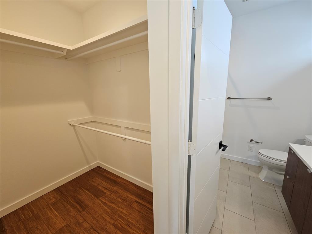 Walk-in closet featuring tiered shelving and hanging rods, wood-finish flooring, and a multi-panel door leading to a bathroom with tile flooring, a vanity with stone-surface countertop, and a wall-mounted towel bar