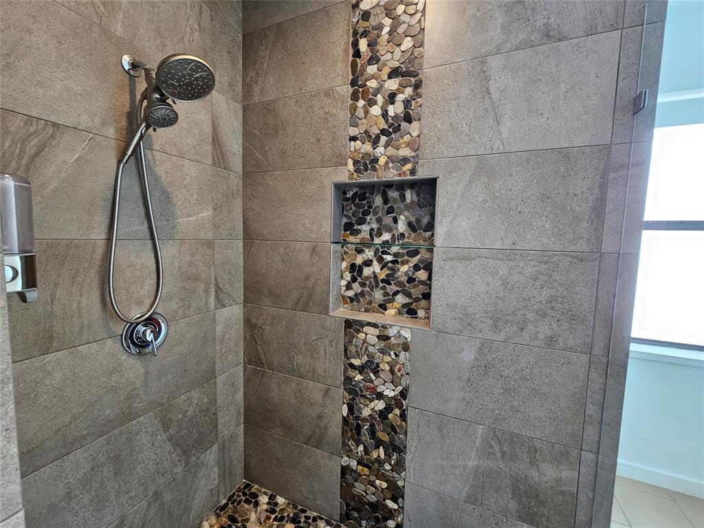 Walk-in shower featuring large-format ceramic tile, a vertical decorative pebble-stone inlay, built-in recessed shelving, a brushed-metal handheld shower fixture, and a natural stone floor