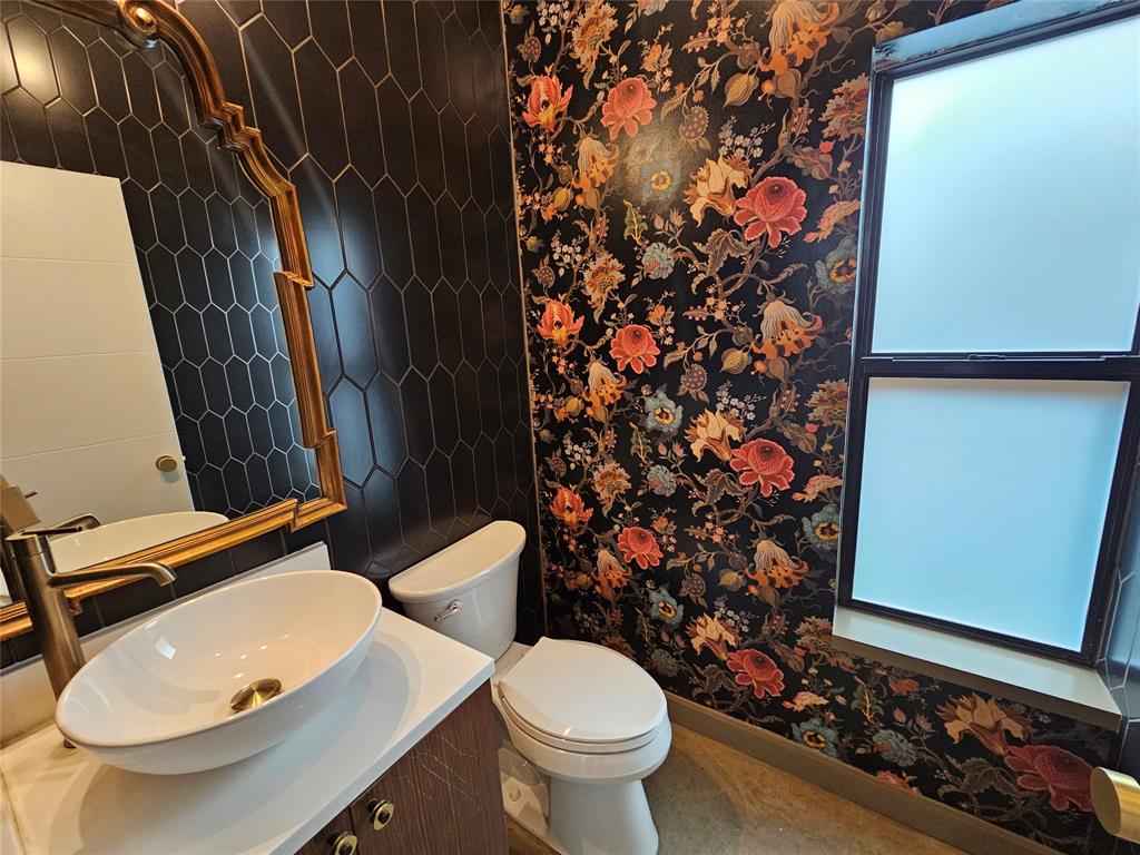 Vessel sink with a brass-finish faucet, black hexagonal tile wall, gold-tone ornate mirror, and floral-patterned wall covering