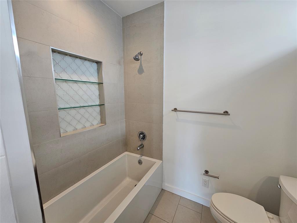 Bathtub with tiled surround, recessed wall niche featuring glass shelving, chrome shower fixtures, and stainless steel hardware