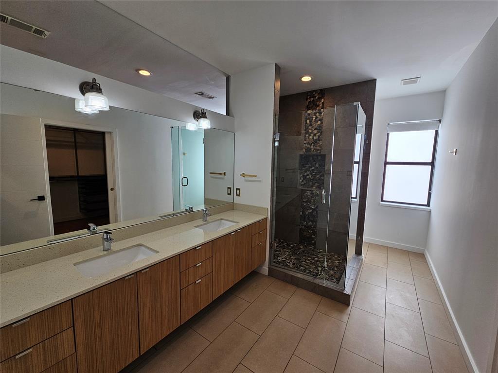 Dual-sink vanity with light-tone countertop, wood-finish cabinetry, full-width wall mirror, and walk-in glass shower featuring an accent tile inlay