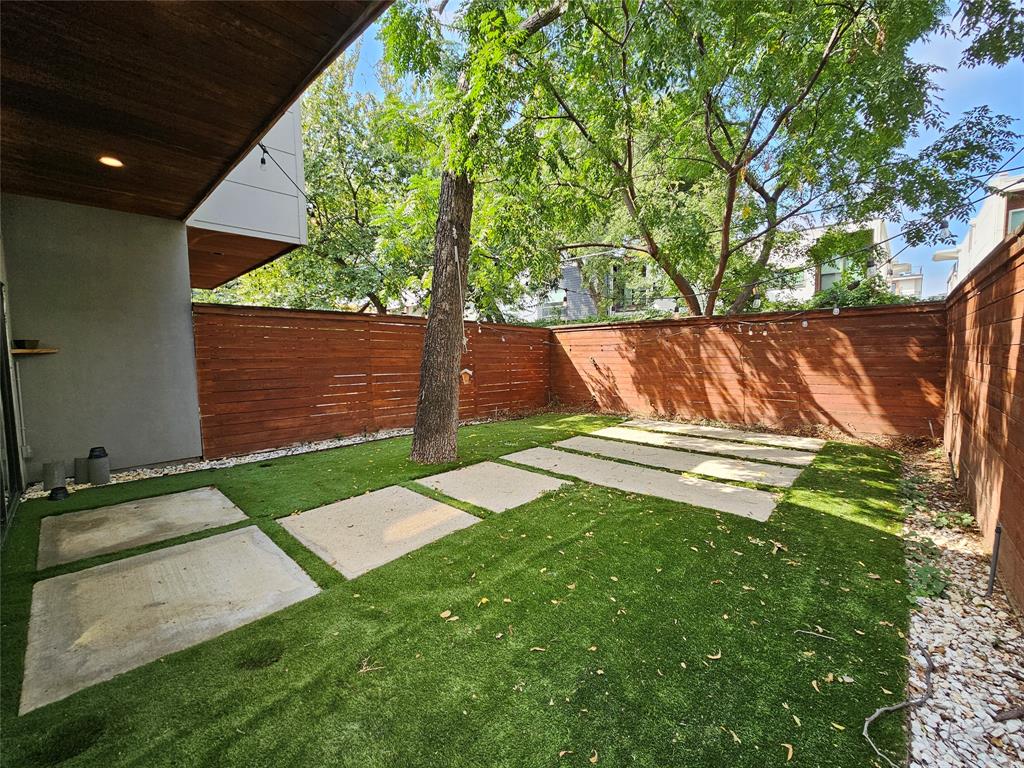 Fenced backyard featuring horizontal wood privacy fencing, artificial turf, concrete pavers, a mature tree, and a covered patio area with a wood-paneled ceiling