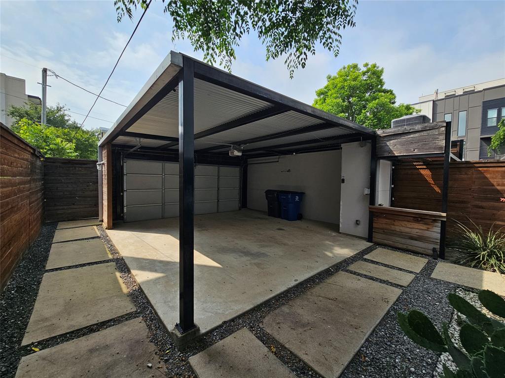 Covered parking structure featuring a steel frame, corrugated metal roof, concrete slab flooring, and integrated wood-accented service area, enclosed by horizontal slat wood fencing and stone-paver hardscaping