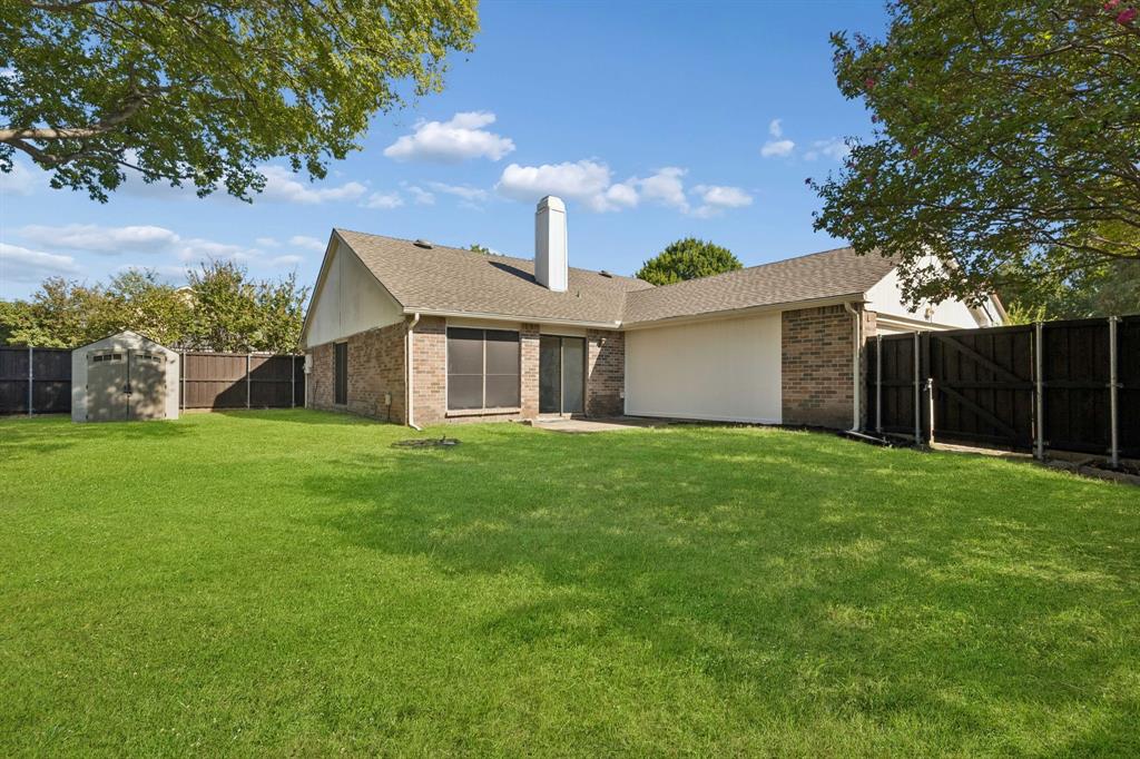 Expansive backyard featuring a manicured lawn, full-height privacy fencing, exterior brick masonry, a central chimney, and a detached storage structure