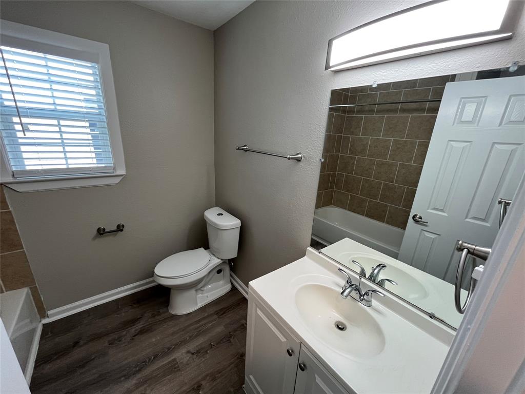 Bathroom featuring a white vanity cabinet, wood-finish flooring, a wall-mounted vanity mirror, a rectangular tub enclosure with tile surround, and brushed nickel hardware