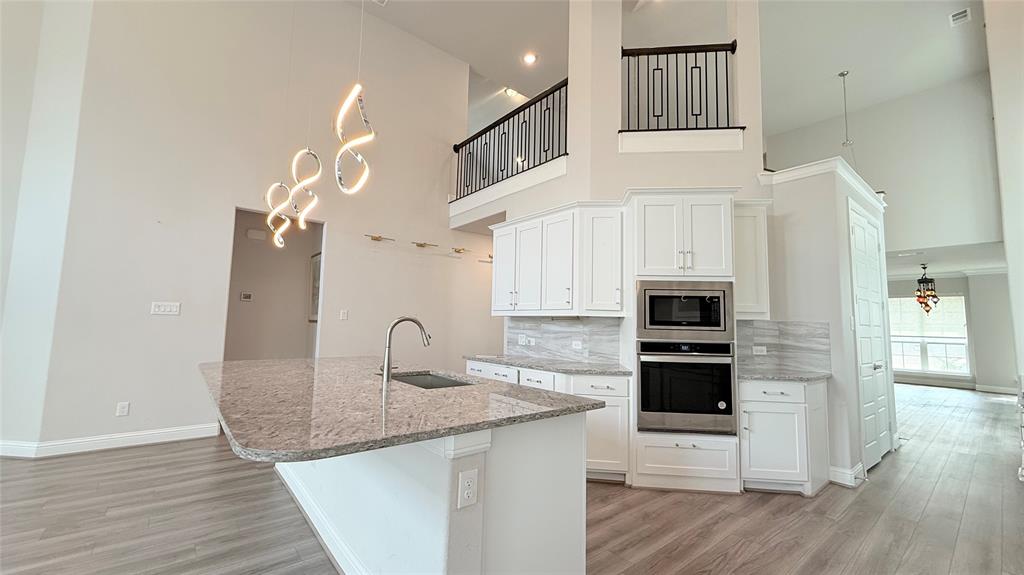 Kitchen featuring high ceilings, white cabinetry, integrated stainless steel double wall ovens, a stone-surface island with a single-basin undermount sink, and wood-finish flooring Kitchen featuring high ceilings, white cabinetry, integrated stainless steel double wall ovens, a stone-surface island with a single-basin undermount sink, and wood-finish flooring