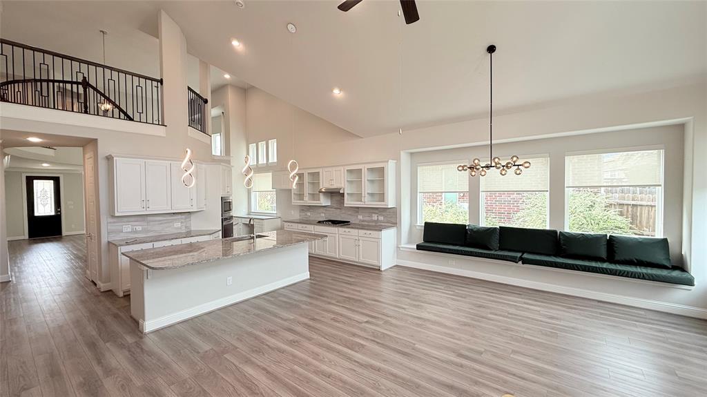 Open-concept kitchen featuring an expansive quartz-topped island, white cabinetry with glass-front accents, wood-finish flooring, vaulted ceilings, and a built-in window bench Open-concept kitchen featuring an expansive quartz-topped island, white cabinetry with glass-front accents, wood-finish flooring, vaulted ceilings, and a built-in window bench