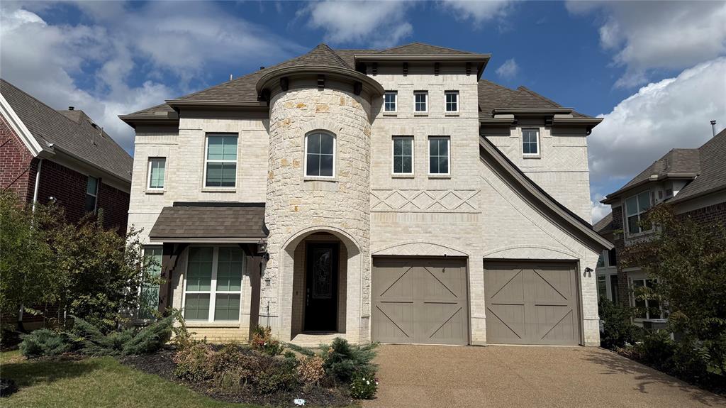 Two-story exterior featuring light-toned brick and stone masonry, a prominent turret entry, decorative masonry detailing, and attached two-car garage with carriage-style doors