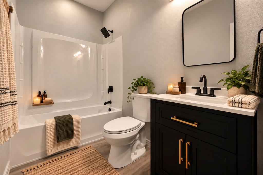 AI INSPIRED -- Bathroom featuring a vanity with a white countertop and gold-toned hardware, a tub-shower combination with matte black fixtures, a rectangular mirror with a rounded black frame, and wood-finish flooring