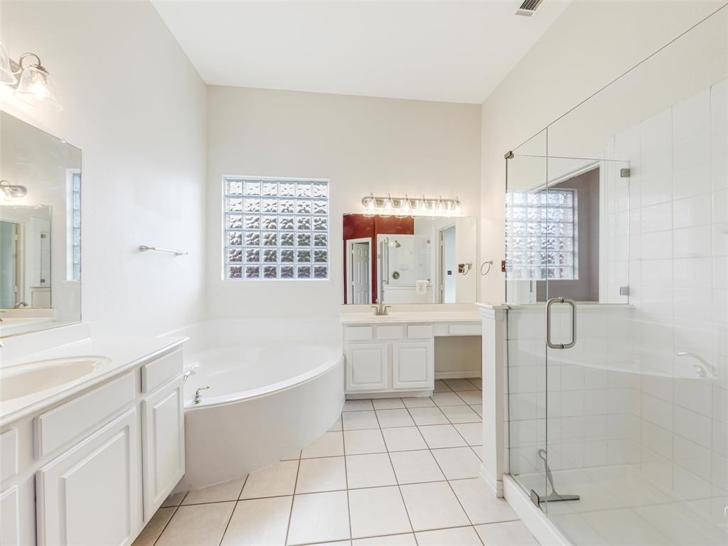 Bathroom featuring a glass-enclosed walk-in shower, a soaking tub, dual-sink vanity with white cabinetry, square glass block window, and ceramic tile flooring