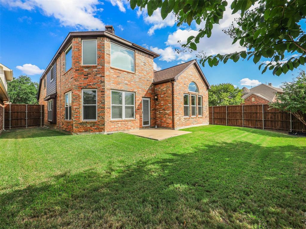 Brick exterior featuring a multi-gabled roofline, expansive lawn, concrete patio, and perimeter wood fencing