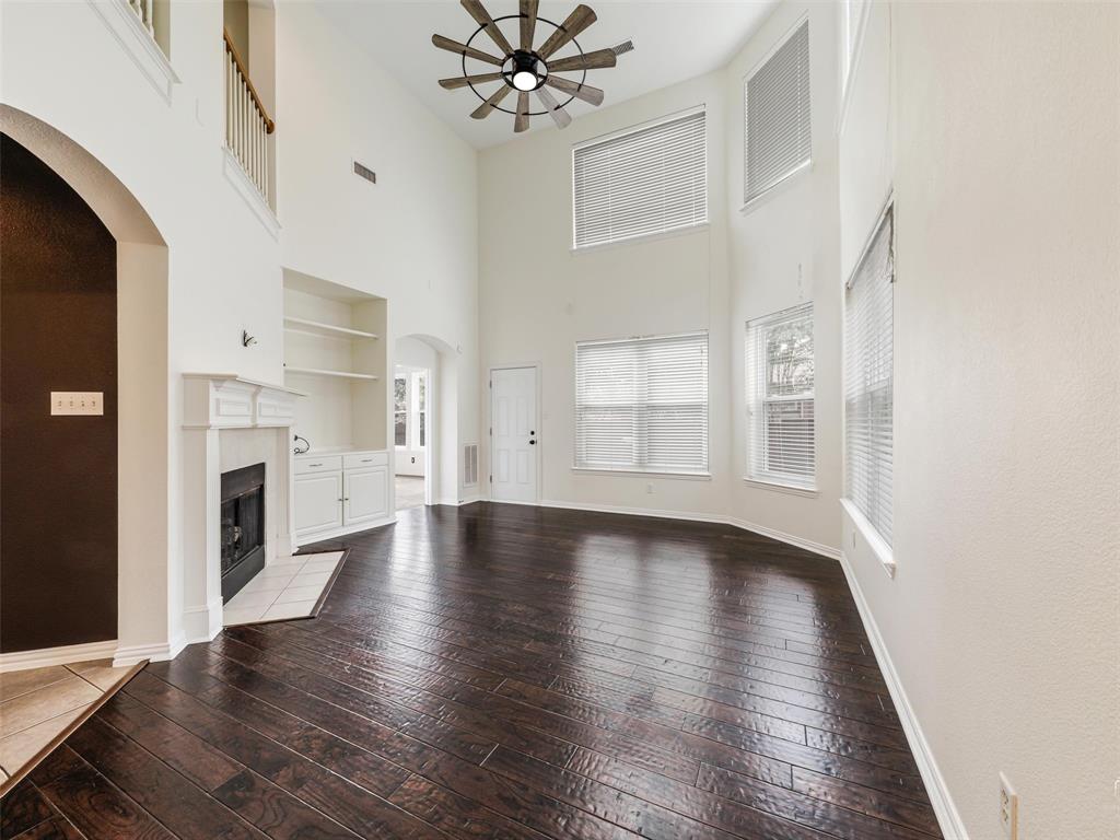 Double-height ceiling featuring a multi-blade ceiling fan, dark wood-finish flooring, built-in cabinetry with shelving, a white fireplace mantel, and large windows