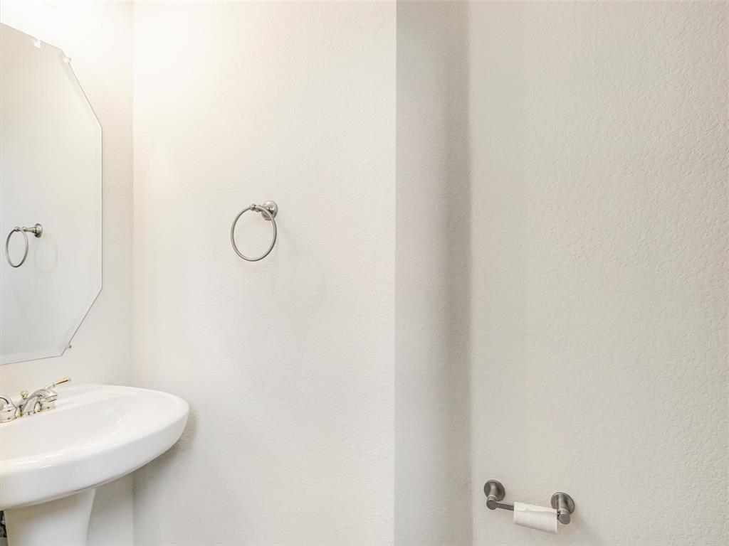 Wall-mounted pedestal sink featuring a polished metal faucet, complemented by an octagonal wall mirror, a circular towel ring, and a coordinating toilet paper holder in a brushed metallic finish