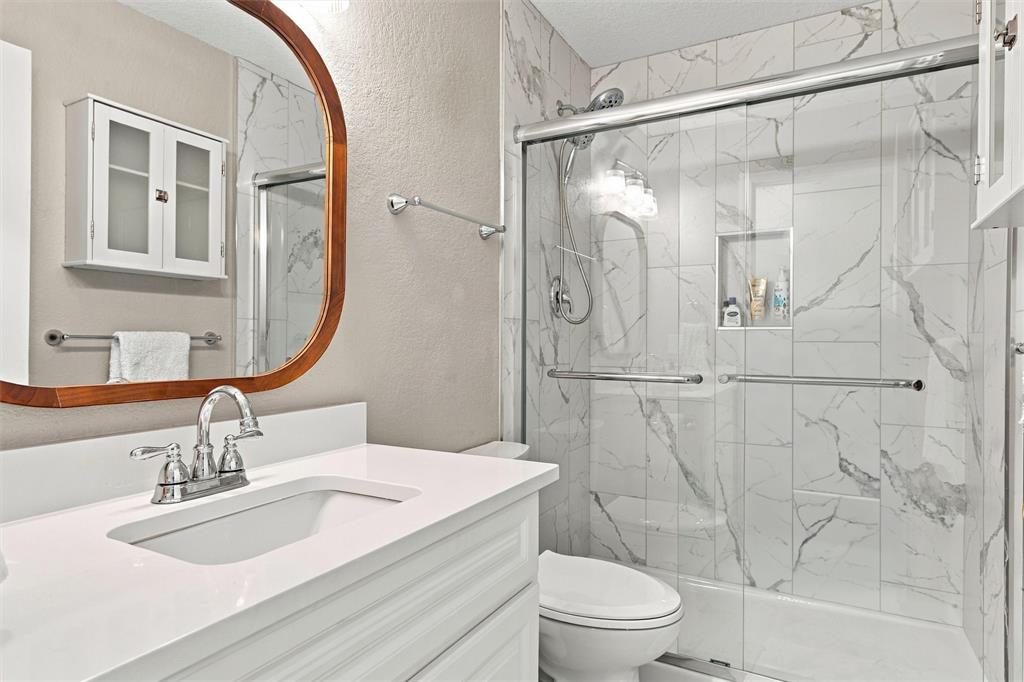 Bathroom featuring a white vanity with a rectangular sink, quartz countertop, polished chrome faucet, arched wood-frame mirror, and shower enclosure with a sliding glass door and integrated tile niche