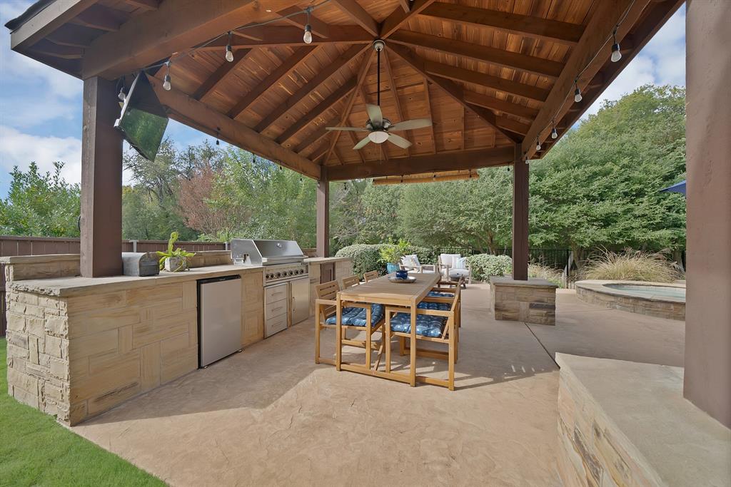 Covered outdoor kitchen featuring a timber-framed pavilion, stone-clad cabinetry, built-in stainless steel grill, beverage refrigerator, and concrete patio flooring