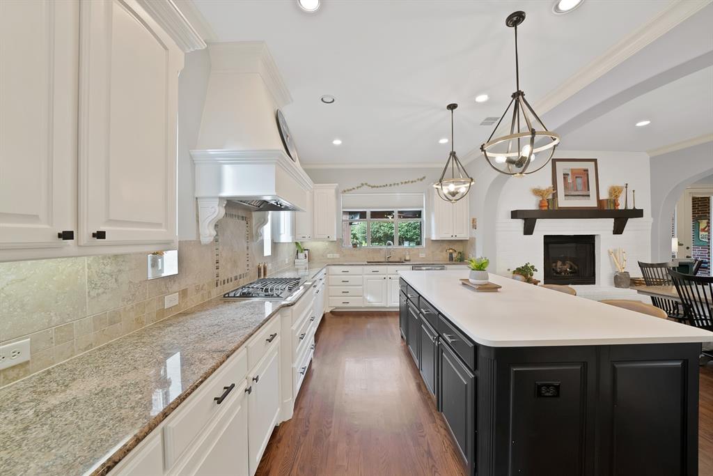 Kitchen featuring wood-finish flooring, a central island with a quartz countertop, stone tile backsplash, white cabinetry with a matching range hood, and a built-in fireplace with a wooden mantel