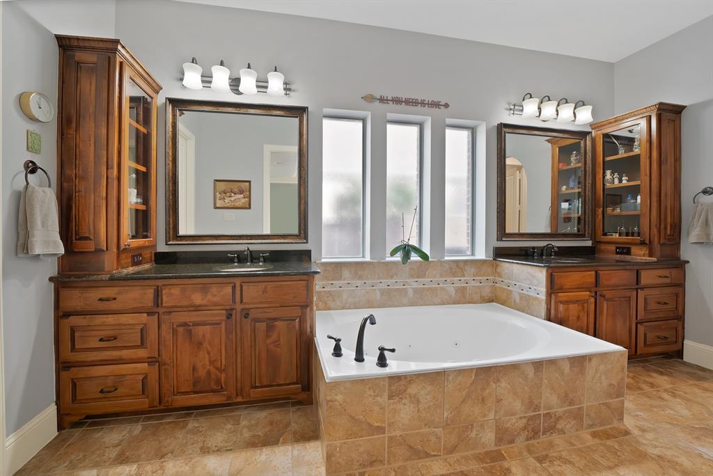 Dual-sink vanity with stained wood cabinetry, dark stone countertops, undermount sinks, tile-surround soaking tub, and matching wall-mounted vanity lighting