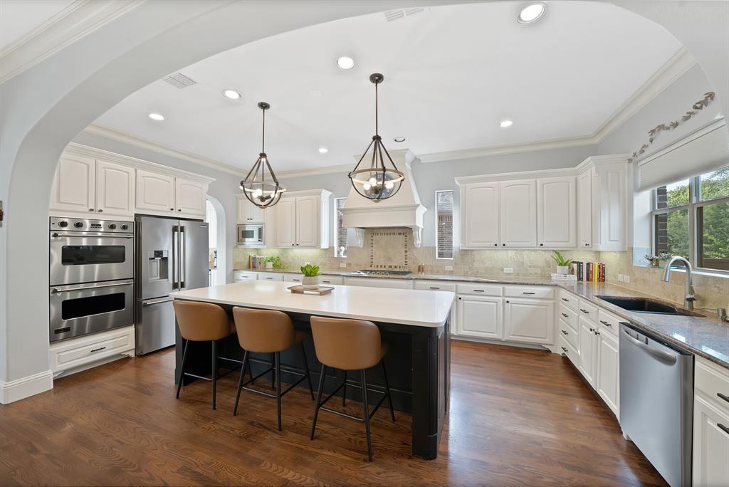 Kitchen featuring white shaker cabinetry, a contrasting dark central island with a solid surface countertop, wood-finish flooring, stainless steel double wall ovens, and custom pendant lighting