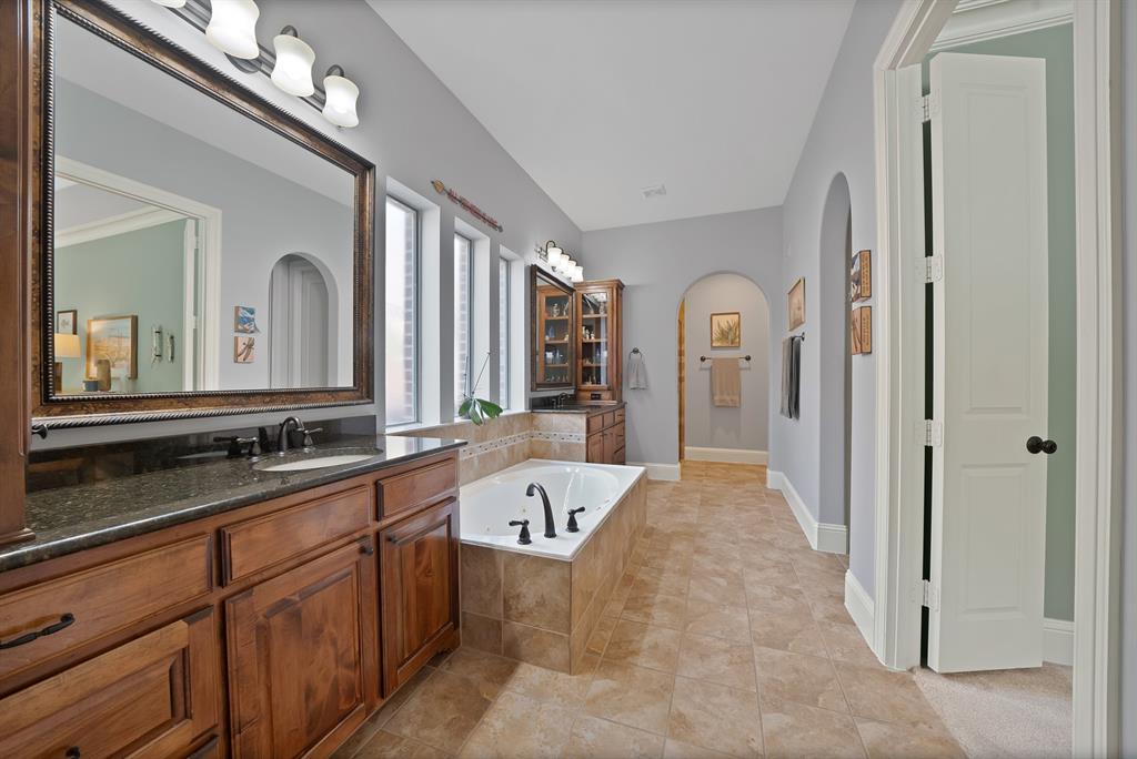 Bathroom featuring wood cabinetry, stone-finish countertops, a drop-in jetted tub with tile surround, ceramic tile flooring, and arched architectural entryways
