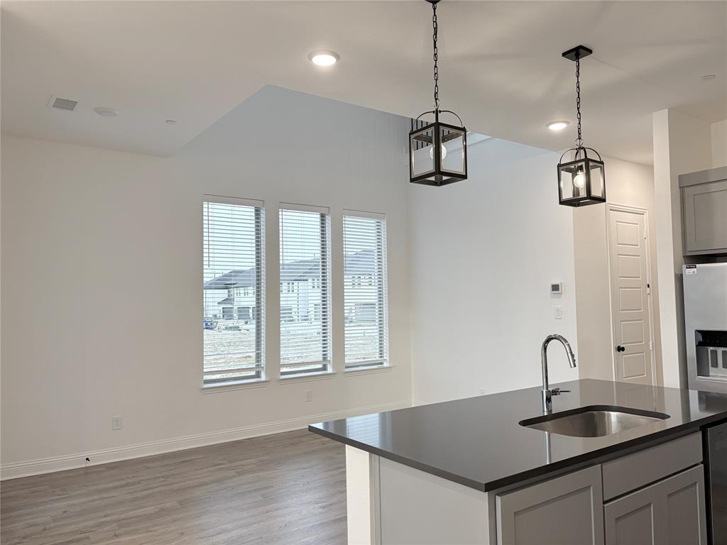 Open-concept kitchen featuring an island with a solid surface countertop, stainless steel undermount sink, shaker-style cabinetry, wood-finish flooring, and pendant lighting Open-concept kitchen featuring an island with a solid surface countertop, stainless steel undermount sink, shaker-style cabinetry, wood-finish flooring, and pendant lighting