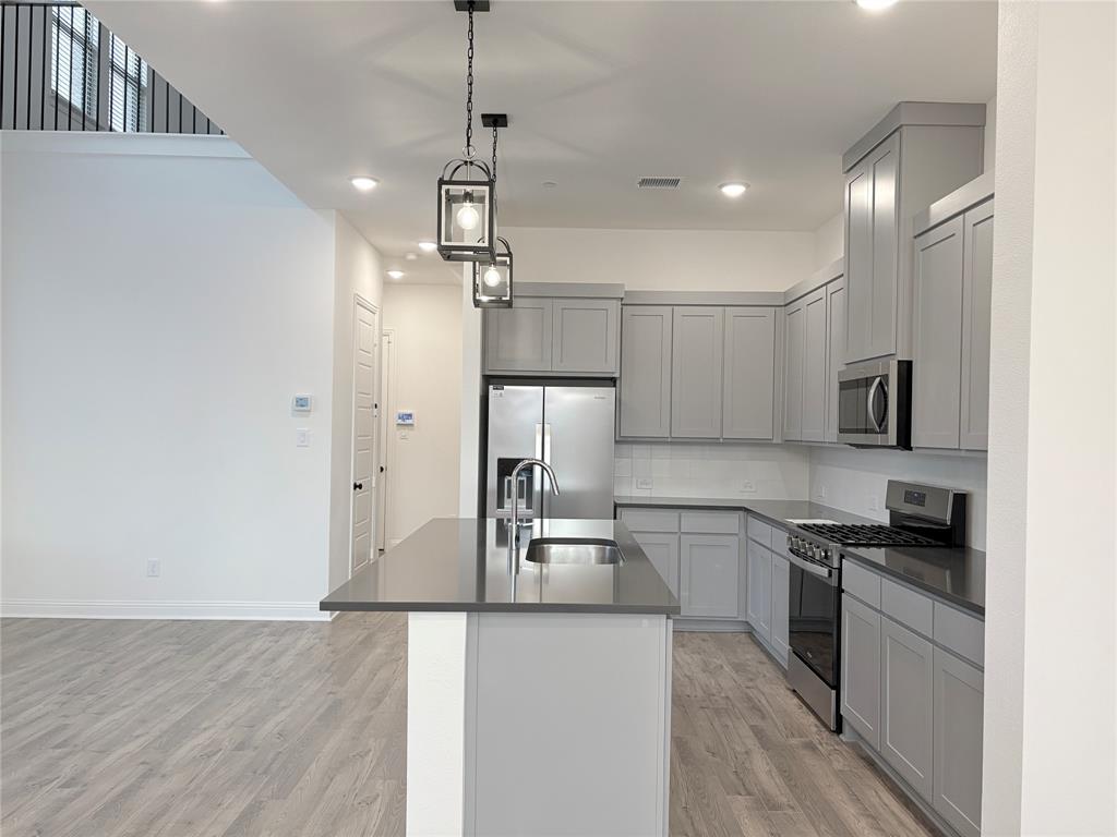 Kitchen featuring Shaker-style cabinetry, a central island with a stainless steel sink, solid-surface countertops, wood-finish flooring, and pendant lighting Kitchen featuring Shaker-style cabinetry, a central island with a stainless steel sink, solid-surface countertops, wood-finish flooring, and pendant lighting