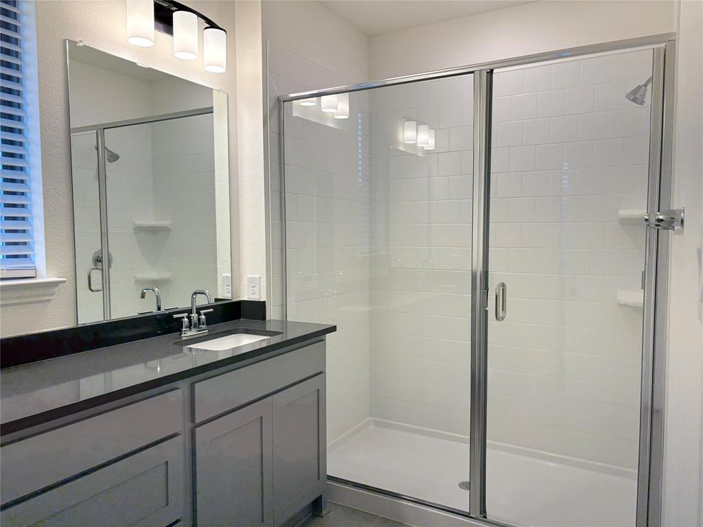 Bathroom featuring a grey vanity with a dark countertop, a walk-in shower with white tile surround and a glass door, a large wall-mounted mirror, and a three-light fixture