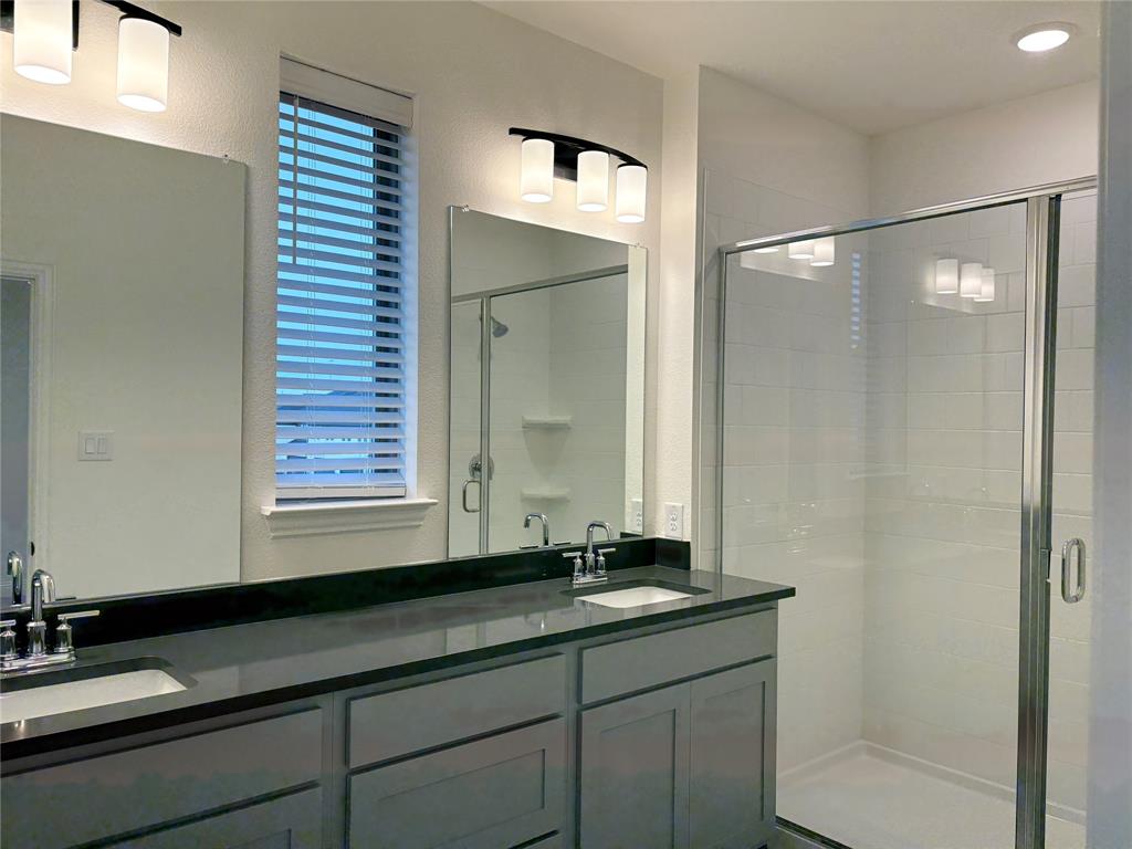 Dual-sink vanity with dark stone-finish countertop, crisp shaker-style cabinetry, glass-enclosed shower, and sleek modern wall-mounted lighting