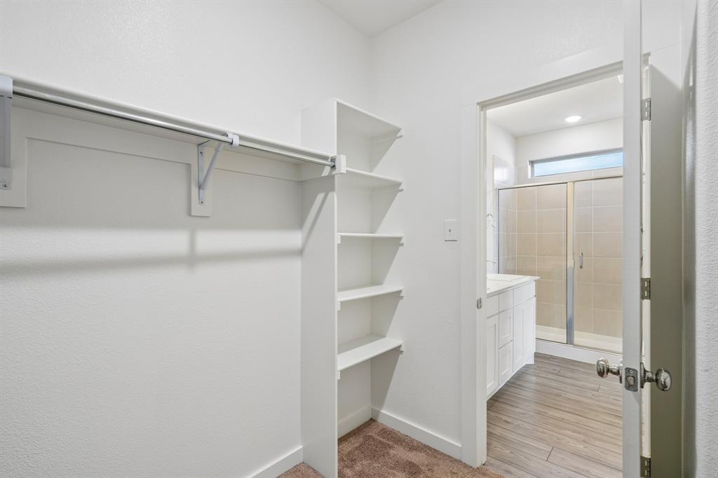 Walk-in closet featuring white built-in shelving and a garment rod, with a doorway revealing a bathroom equipped with wood-finish flooring, a white cabinet vanity, and a tiled shower stall
