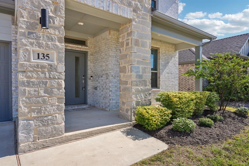 Covered front porch featuring stacked stone accents, gray-toned masonry, a modern entry door with a vertical glass insert, and wall-mounted exterior cylinder lighting