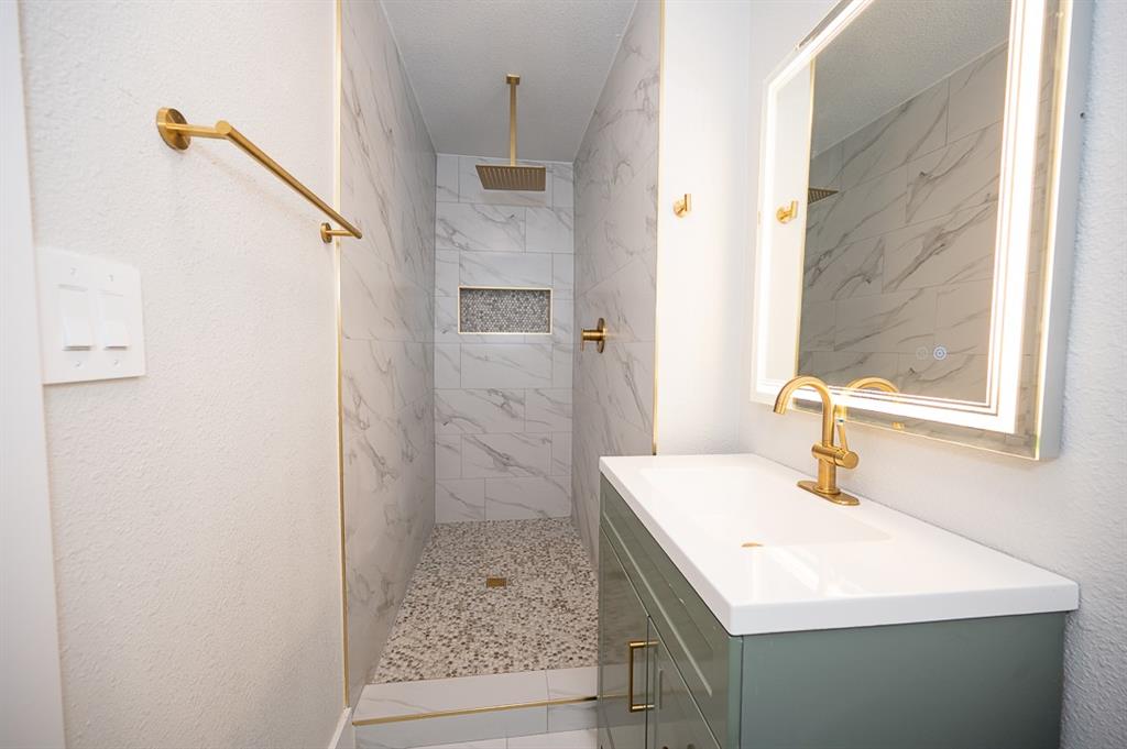 Walk-in shower featuring tiled walls with stone-pattern detail, pebble-tile flooring, and a rainfall showerhead; vanity with a white countertop and gold-finish hardware; backlit mirror